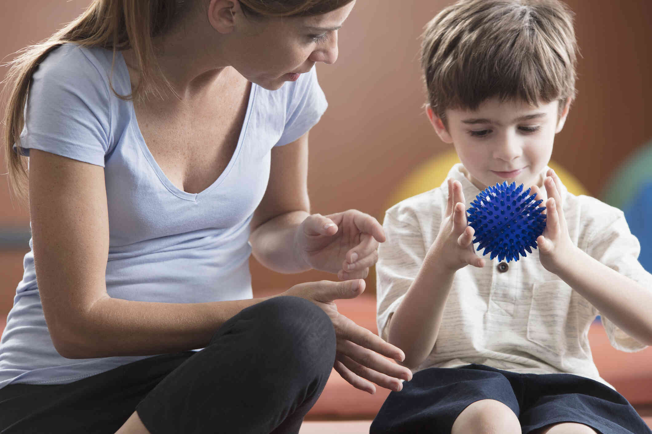 A young boy holds a blue ball in his hands with a smile as a woman sits next to him and smiles at him.